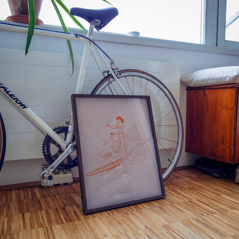 May include: A framed print featuring an orange line drawing of two people surfing. The artwork is in a dark brown frame and leans against a white bicycle. The background includes a wooden cabinet and a window.