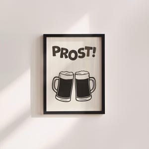 May include: Black framed print with the word "PROST!" above two beer mugs. The mugs are filled with dark liquid and topped with white foam. The artwork is a simple, black and white illustration, suitable for a kitchen or bar.