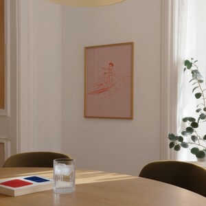 May include: A framed pink print featuring a surfer and child on a surfboard. The artwork has a light brown frame and is hanging on a white wall. A glass of water with ice sits on a wooden table.