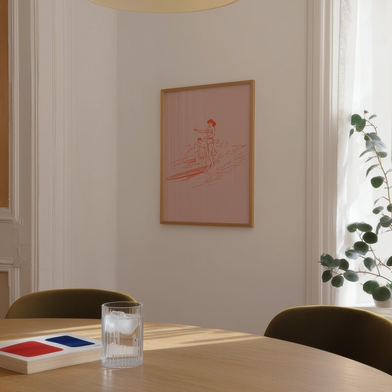 May include: A framed pink print featuring a surfer and child on a surfboard. The artwork has a light brown frame and is hanging on a white wall. A glass of water with ice sits on a wooden table.