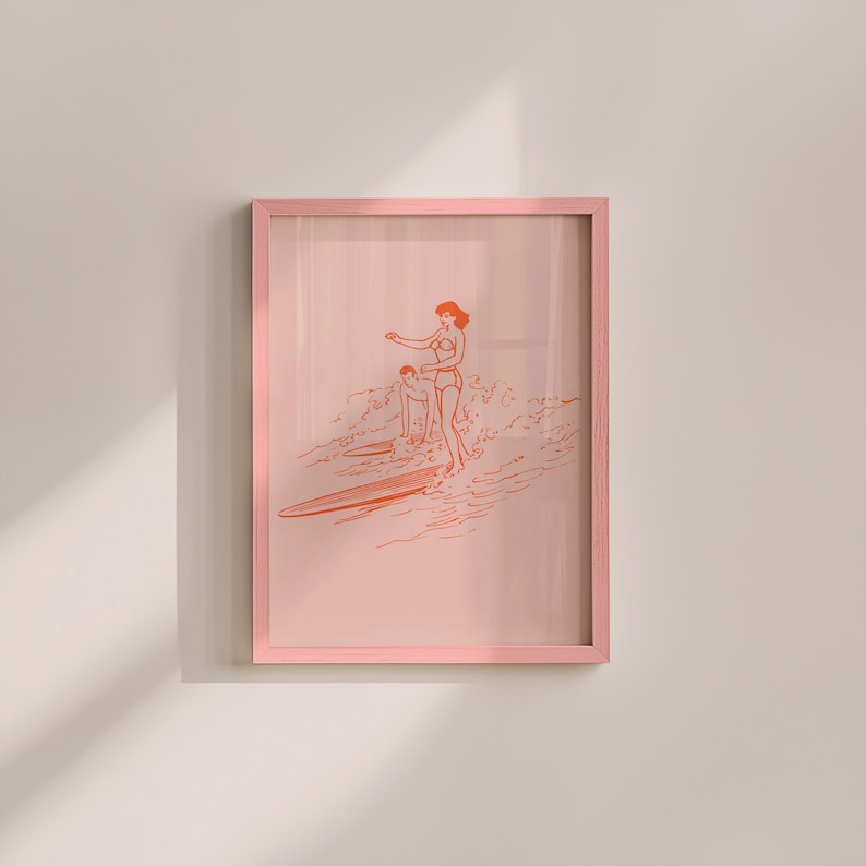 May include: A framed art print featuring a line drawing of two people surfing. The artwork is in shades of orange and pink, with a pink frame. The image depicts a woman and a child on surfboards in the ocean.