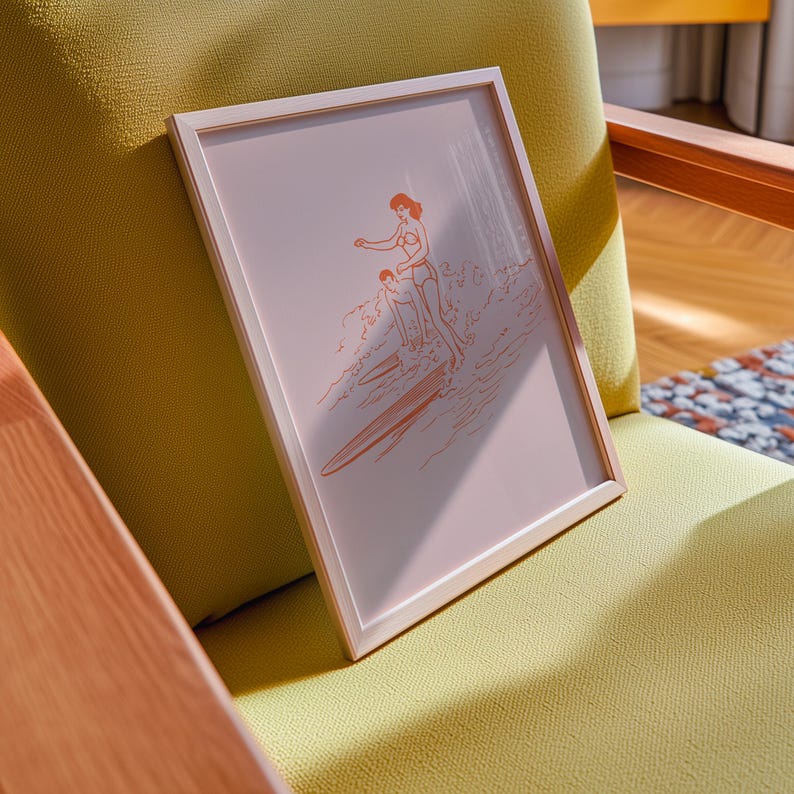 May include: A framed print featuring an orange line drawing of two people surfing. The artwork is set against a pale pink background and framed in a light wood-tone frame. The print is resting on a yellow chair.