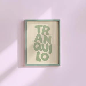 May include: A framed art print featuring the word "TRANQUILO" in green, bubble-style lettering. The print has a cream-coloured background and is encased in a muted green frame. The artwork is displayed on a light purple wall.