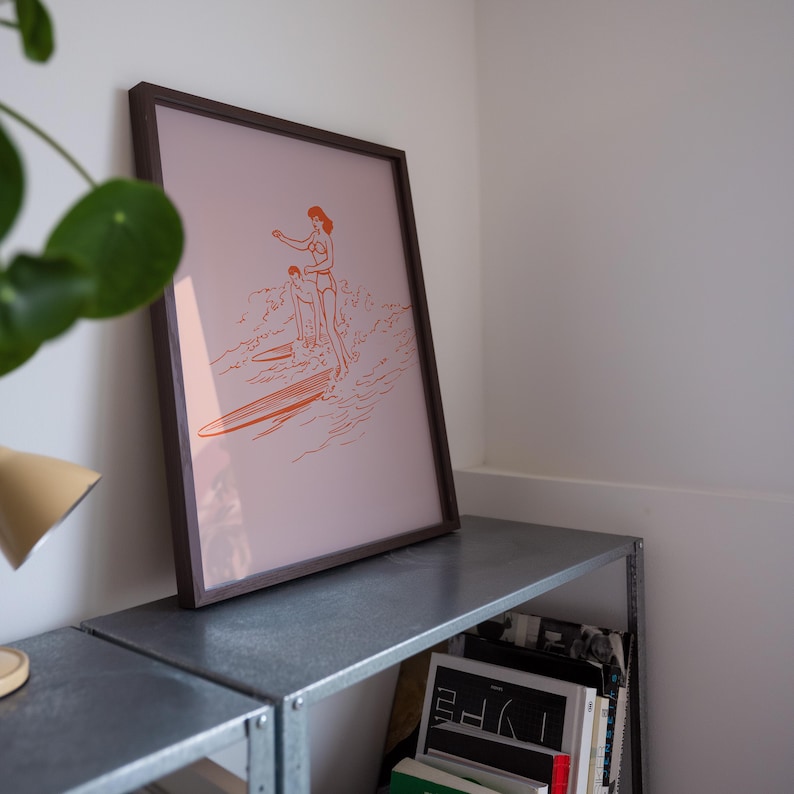 May include: Framed art print featuring an orange line drawing of two people surfing on a wave. The artwork is set against a pale pink background and framed in a dark brown wooden frame. The print is displayed on a metal shelf.