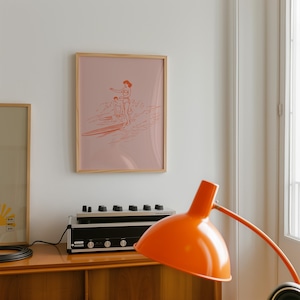 May include: A framed print featuring a minimalist illustration of surfers on a wave in shades of coral on a pink background. The artwork is encased in a light wood frame, adding a touch of warmth to the piece. The print is displayed on a white wall.