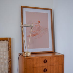 May include: A framed art print featuring a minimalist illustration of two people surfing, in shades of orange and pink. The print is displayed on a wooden dresser, next to a gold-toned desk lamp.