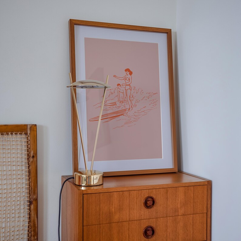 May include: A framed art print featuring a minimalist illustration of two people surfing, in shades of orange and pink. The print is displayed on a wooden dresser, next to a gold-toned desk lamp.
