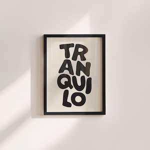 May include: A framed art print featuring the word "TRANQUILO" in large, black, rounded letters. The text is set against a cream background, within a black frame. The artwork is mounted on a white wall.