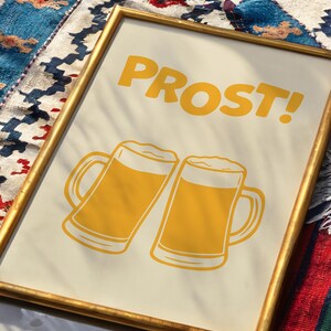 May include: A framed print with a gold frame and a white background. The print features two yellow cartoon beer mugs with the word "PROST!" in yellow letters.