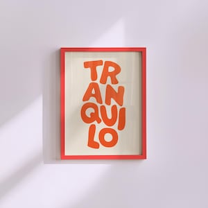 May include: A framed art print with the word "TRANQUILO" in orange, bold, rounded letters. The print is set against a cream background and framed in a bright red frame. The artwork is displayed on a white wall.