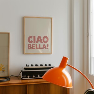 May include: A framed print with the words "CIAO BELLA!" in pink letters hangs on a white wall. An orange desk lamp and a stereo system sit on a wooden cabinet below the print. The room has a modern, minimalist aesthetic.