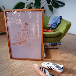 May include: A framed print with a surfing theme. The artwork features an orange line drawing of a person surfing. The frame is made of wood. A green chair, blue jeans, a red book, and white sneakers with blue stripes are also visible.