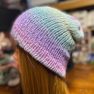 May include: A hand-knitted beanie hat featuring a variegated yarn pattern with blended shades of pastel purple, soft pink, and light mint green. The knit texture displays a consistent ribbed stitch pattern throughout the entire head covering.