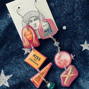May include: Dangle earrings in pink and silver, with images of a woman, a tombstone, a moon, and a cross. The earrings also feature the text "Kiss the Librarian" and "Sunnydale".