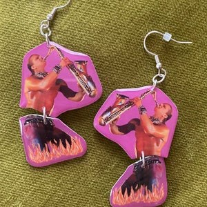 May include: A pair of pink resin earrings featuring a man playing a saxophone with flames on the bottom.