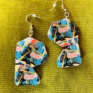 May include: A pair of earrings made from comic book pages. The earrings are shaped like a jagged, irregular hexagon. The comic book pages feature a superhero in a blue and red costume riding a motorcycle. The text on the comic book pages reads "BLOODIEST ISSUE EVER!" and "DESTROY ALL".