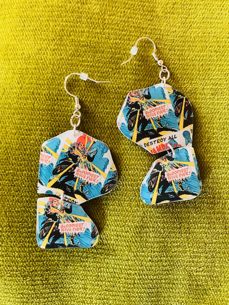 May include: A pair of earrings made from comic book pages. The earrings are shaped like a jagged, irregular hexagon. The comic book pages feature a superhero in a blue and red costume riding a motorcycle. The text on the comic book pages reads "BLOODIEST ISSUE EVER!" and "DESTROY ALL".