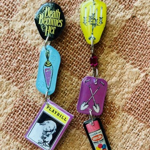 Puede incluir: Un par de pendientes colgantes con varios dijes. Un pendiente presenta un dije negro con el texto "Death Becomes Her", un dije de poción azul y un programa. El otro pendiente tiene un dije de candelabro amarillo, un dije de pala morada y un dije de pintura en aerosol Krylon.