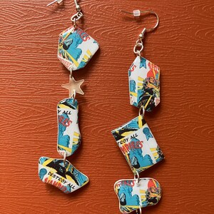 Peut inclure: Une paire de boucles d'oreilles longues et pendantes r&eacute;alis&eacute;es &agrave; partir de pages de bandes dessin&eacute;es recycl&eacute;es. Les boucles d'oreilles pr&eacute;sentent un charme d'&eacute;toile argent&eacute;e et un charme de croix argent&eacute;e. Les pages de bandes dessin&eacute;es pr&eacute;sentent des images de monstres et le texte "Destroy All Vampires".