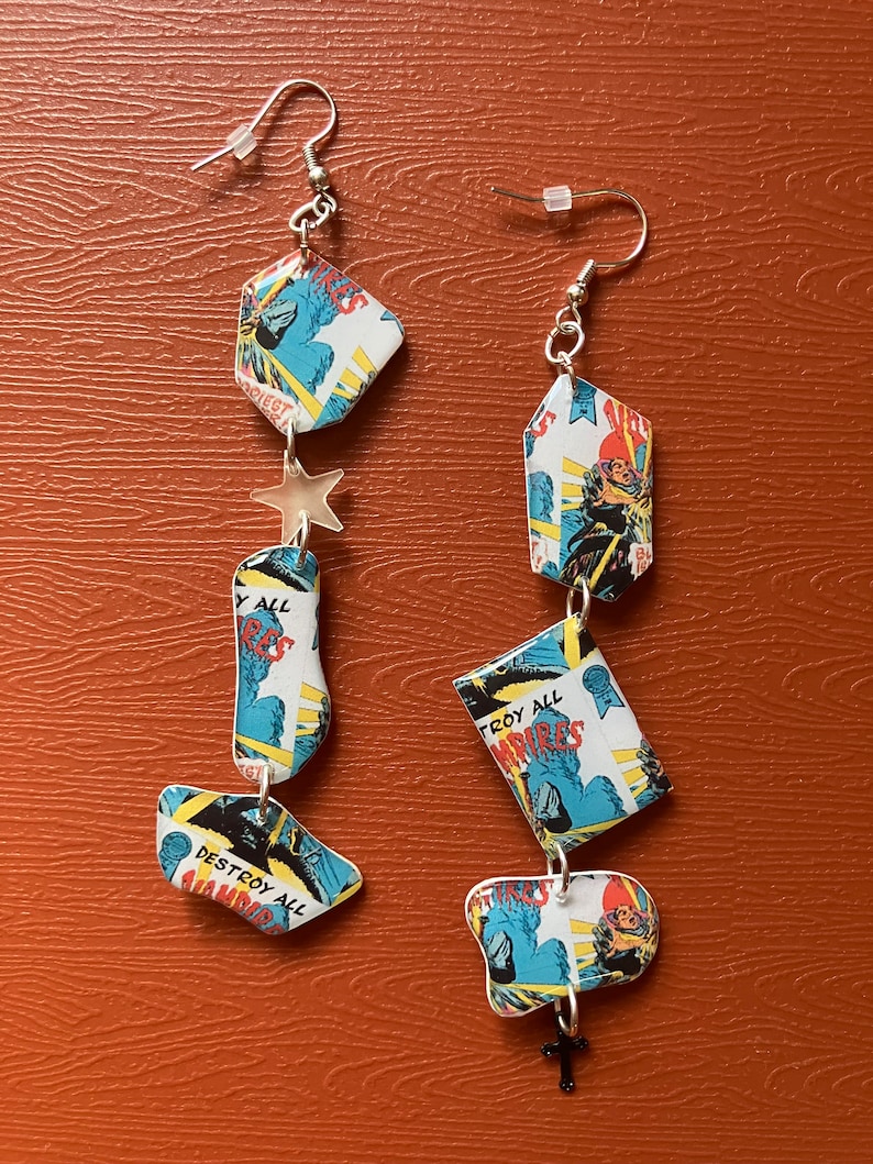 Peut inclure: Une paire de boucles d'oreilles longues et pendantes r&eacute;alis&eacute;es &agrave; partir de pages de bandes dessin&eacute;es recycl&eacute;es. Les boucles d'oreilles pr&eacute;sentent un charme d'&eacute;toile argent&eacute;e et un charme de croix argent&eacute;e. Les pages de bandes dessin&eacute;es pr&eacute;sentent des images de monstres et le texte "Destroy All Vampires".