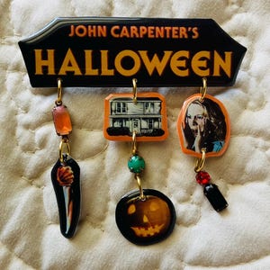 May include: A black and orange Halloween-themed brooch featuring the text "JOHN CARPENTER'S HALLOWEEN." The brooch has dangling charms including a knife, a house, a jack-o'-lantern, and a woman's face. The charms are connected with gold-colored hoops and beads.