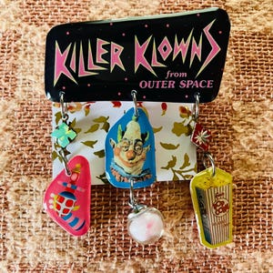 Può includere: Un cartello nero con le parole "KILLER KLOWNS from OUTER SPACE" in lettere rosa. Sotto il cartello ci sono diversi ciondoli pendenti, tra cui una forma rosa con un disegno, una faccia da clown, una palla trasparente e una scatola di popcorn.