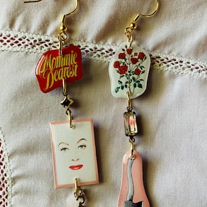 Op de afbeelding: Een paar gouden oorhangers met een rood, wit en roze thema. De oorbellen hebben een rode hanger met de tekst "Mommie Dearest", een witte hanger met een bloemenontwerp, een heldere kristallen hanger, een roze hanger met een bijl en een gouden hanger in de vorm van een kleerhanger.