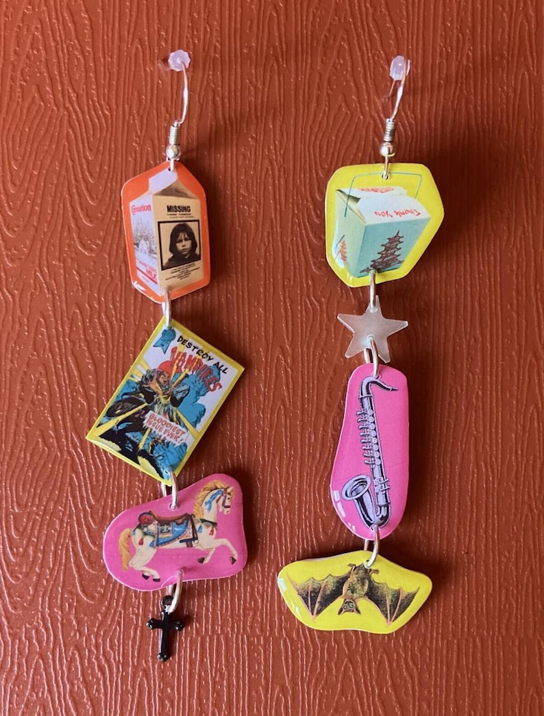 Peut inclure: Une paire de boucles d'oreilles pendantes avec une vari&eacute;t&eacute; de breloques. Les breloques comprennent un carton de lait avec une affiche d'enfant disparu, une bo&icirc;te &agrave; emporter chinoise, une &eacute;toile, un saxophone, un cheval de carrousel et une chauve-souris.