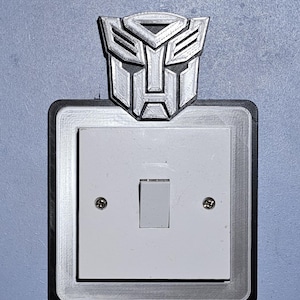 May include: A white light switch cover featuring a silver Transformers logo. The cover is square with a silver border, and a white switch is in the centre. The background is a light blue wall.