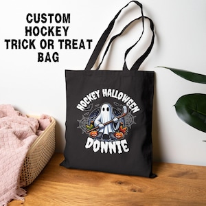 K&ouml;nnte beinhalten: Eine schwarze Tragetasche mit einer Grafik eines Geistes, der Schlittschuhe tr&auml;gt und einen Hockeyschl&auml;ger h&auml;lt, umgeben von K&uuml;rbissen und Spinnenweben. Der Text lautet HOCKEY HALLOWEEN DONNIE. Die Tasche hat zwei schwarze Schulterriemen.