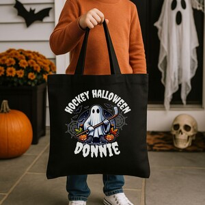 K&ouml;nnte beinhalten: Eine schwarze Tragetasche mit einer Grafik eines Geistes, der Schlittschuhe tr&auml;gt und einen Hockeyschl&auml;ger h&auml;lt. Das Design zeigt orangefarbene K&uuml;rbisse, Spinnweben und Flederm&auml;use sowie den wei&szlig;en Schriftzug HOCKEY HALLOWEEN DONNIE in Bogenform.