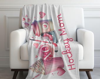 May include: White throw blanket with a watercolor design featuring hockey equipment, a coffee cup, and a dessert cup with sprinkles. The words "Hockey Mom" are written vertically in pink. Pink hearts are scattered across the blanket.
