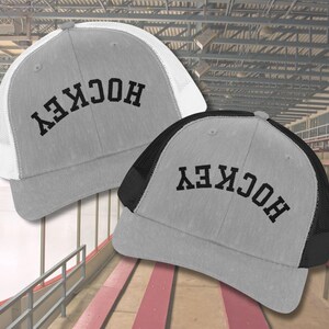 May include: Two grey and black mesh trucker hats with the word "HOCKEY" embroidered in black on the front. The hats have a white mesh back on one and a black mesh back on the other.