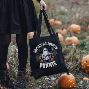 K&ouml;nnte beinhalten: Eine schwarze Tragetasche aus Segeltuch mit einer Grafik eines Geistes, der Schlittschuhe tr&auml;gt und einen Hockeyschl&auml;ger h&auml;lt. Der Text lautet HOCKEY HALLOWEEN DONNIE, umgeben von Illustrationen aus Herbstbl&auml;ttern, K&uuml;rbissen und Spinnenweben.