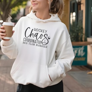 May include: Cream-coloured hoodie with the text "Hockey Chaos Coordinator AKA Team Manager" in black script. The hoodie has a front pocket and a drawstring hood. A coffee cup is held in the hand.