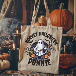 K&ouml;nnte beinhalten: Eine beige Tragetasche aus Segeltuch mit einer Grafik eines Geistes, der Schlittschuhe tr&auml;gt und einen Hockeyschl&auml;ger h&auml;lt. Der Text HOCKEY HALLOWEEN DONNIE ist von Spinnweben, Flederm&auml;usen und K&uuml;rbissen umgeben, gedruckt in Schwarz und Orange.