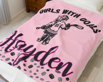 May include: Pink blanket with a hockey-themed design. Features the text "GIRLS WITH GOALS" and the name "Hayden" in black script. Includes a black and white illustration of a hockey player and hockey pucks.