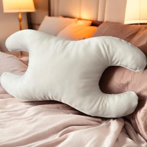 Könnte beinhalten: Ein weißes, konturiertes Kissen für den Komfort in Bauchlage. Das Kissen hat eine einzigartige Form mit gebogenen Kanten und einem zentralen Ausschnitt. Es liegt auf einem Bett mit rosa und weißer Bettwäsche. Die glatte Oberfläche deutet auf ein weiches, atmungsaktives Material hin.