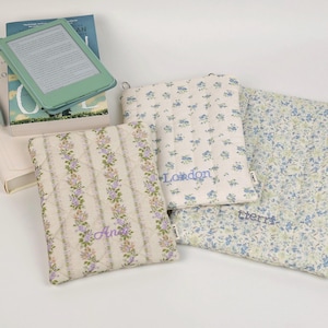 May include: Three quilted fabric sleeves for electronic devices featuring floral patterns in blue, green, and purple. Each sleeve is embroidered with a name: Ana, London, and tierri. The sleeves are displayed alongside a book and an e-reader.