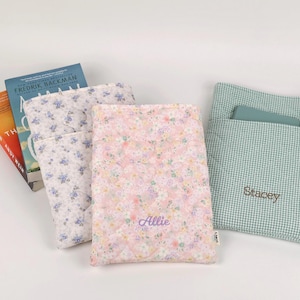May include: Three rectangular fabric book sleeves with quilted textures. One features a blue floral pattern, one a pink floral pattern with the name Allie embroidered in purple, and one a green gingham check with the name Stacey embroidered in brown.