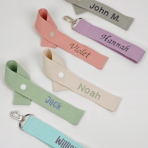 May include: Five fabric keychains in sage green, dusty pink, cream, light green, and turquoise. Each features a snap button and embroidered names: John M., Hannah, Violet, Noah, Jack, and William. The straps include silver metal clasps.