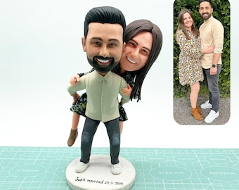 Op de afbeelding: Een gepersonaliseerd bobblehead-beeldje van een stel, de man in een lichtgroen overhemd en donkere jeans, de vrouw omhelst hem van achteren. De basis luidt "Just married 29.11.2024". Een foto van het stel staat op de achtergrond.