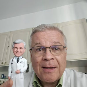 May include: A man wearing glasses and a white lab coat is standing in front of a white cabinet. He is smiling and looking at the camera. A small figurine of a man wearing a white lab coat and a stethoscope is standing in front of him.