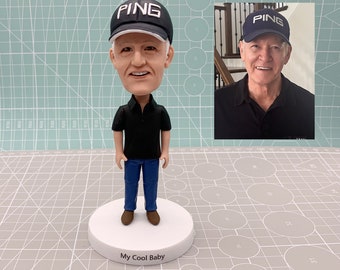 May include: A black and blue bobblehead figurine of a person wearing a black shirt, blue jeans, and a black baseball cap with the word "PING" in white lettering. The bobblehead is standing on a white base with the words "My Cool Baby" printed on it.