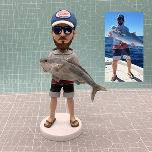 May include: A figurine of a person holding a large fish. The figure wears a gray hoodie, black shorts, and a blue cap with text. The fish is gray and detailed. The figurine stands on a white base. An inset photo shows a person holding a fish.