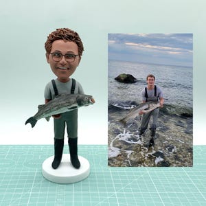 May include: A figurine of a person wearing glasses and fishing attire, holding a striped fish. The figurine is on a white base. A photograph of a person holding a fish in the ocean is in the background.