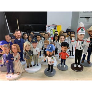 May include: A collection of bobblehead figurines, each depicting a person in various outfits. The figurines are on round bases and include figures in medical attire, formal wear, and casual clothing. The bobbleheads are arranged on a light-colored surface.