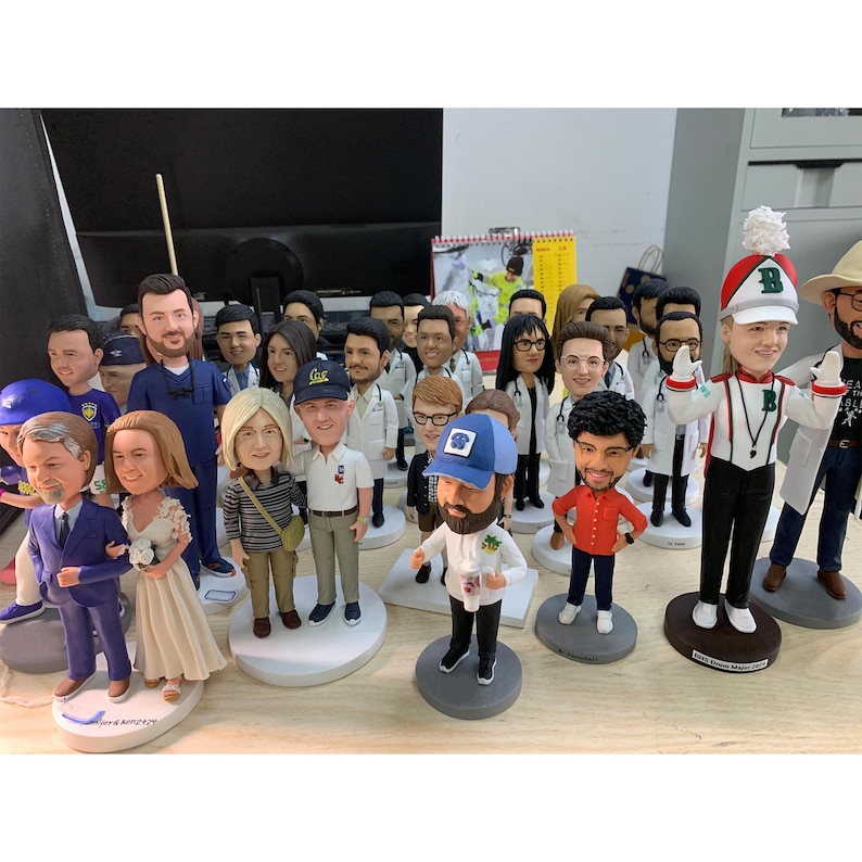 May include: A collection of bobblehead figurines, each depicting a person in various outfits. The figurines are on round bases and include figures in medical attire, formal wear, and casual clothing. The bobbleheads are arranged on a light-colored surface.