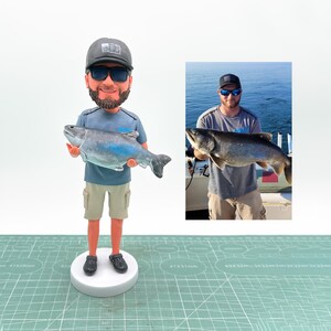 May include: A figurine of a man holding a large fish, with a smaller photo of the man holding the fish. The figurine wears a gray cap, sunglasses, a blue shirt, khaki shorts, and black shoes. The fish is gray and blue.