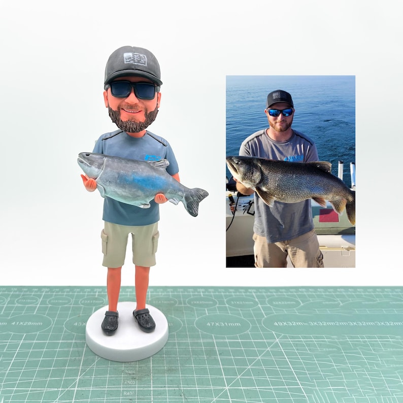 May include: A figurine of a man holding a large fish, with a smaller photo of the man holding the fish. The figurine wears a gray cap, sunglasses, a blue shirt, khaki shorts, and black shoes. The fish is gray and blue.